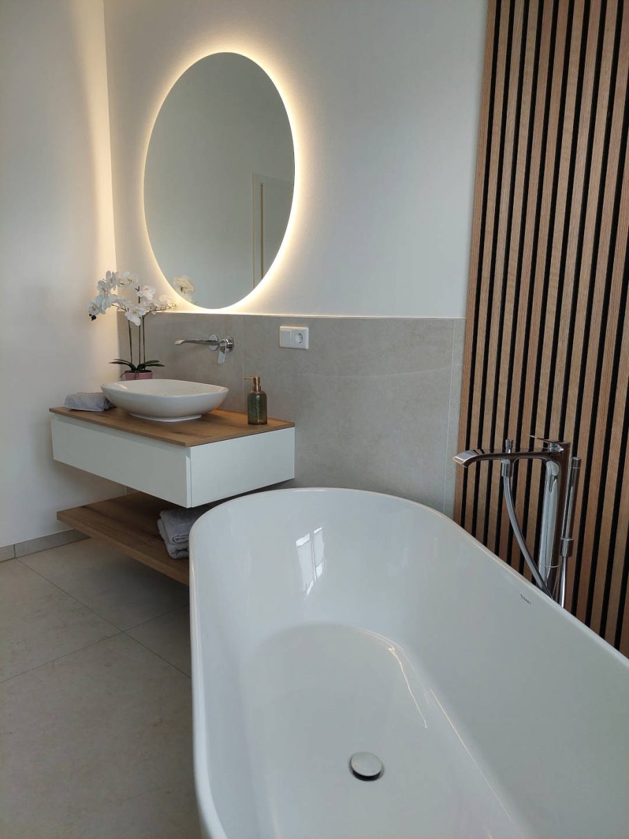Moderne Badgestaltung mit runder Spiegelleuchte, freistehender Badewanne und Holzelementen.