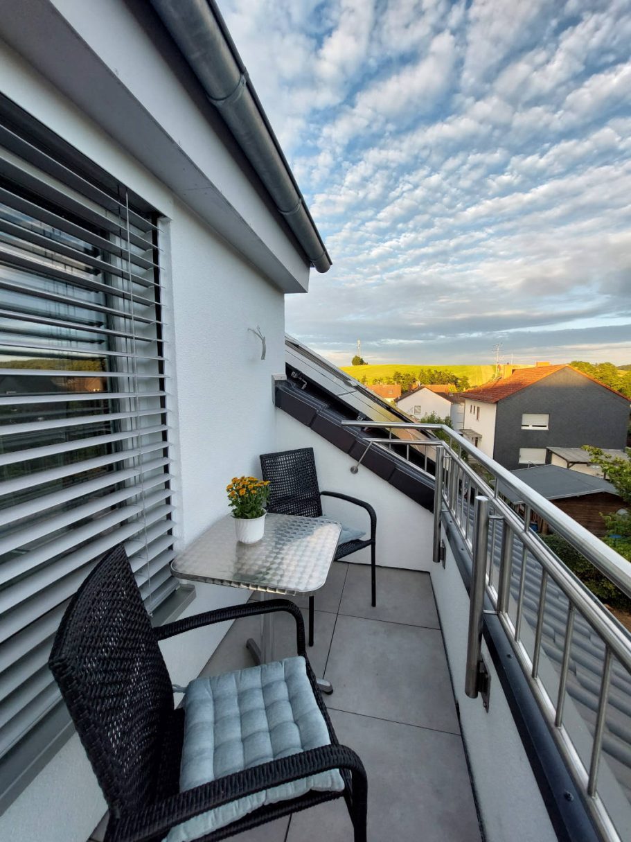 Balkon mit zwei Stühlen, Tisch und Pflanzen, wolkiger Himmel im Hintergrund.